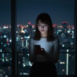 試験の翌日、スマートフォンにSMSで届いた俳協ボイスの不合格通知を見て、ショックを受けて立ち尽くす若いアジア人女性。都会の夜景を背景に、不安な心境を表す